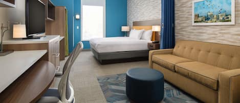 A hotel room with a bed, a sofa, a desk with a chair, a TV, and a painting on the wall.