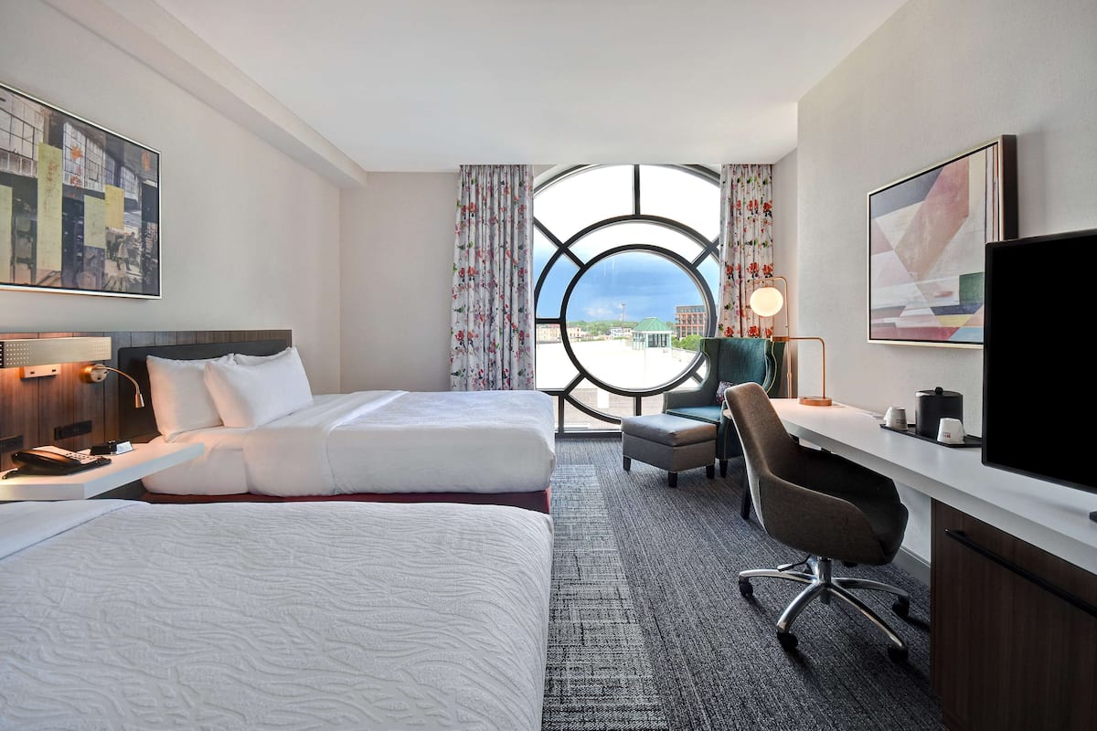 A hotel room with two beds, a desk, a chair, a TV, and a large window with a view of buildings.