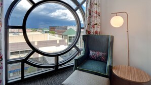 Ein Zimmer mit einem großen Rundfenster, das einen Blick auf Gebäude und ein Gebäude mit Gründach bietet. Ein Sessel mit Kissen und ein kleiner Tisch befinden sich in der Nähe des Fensters.