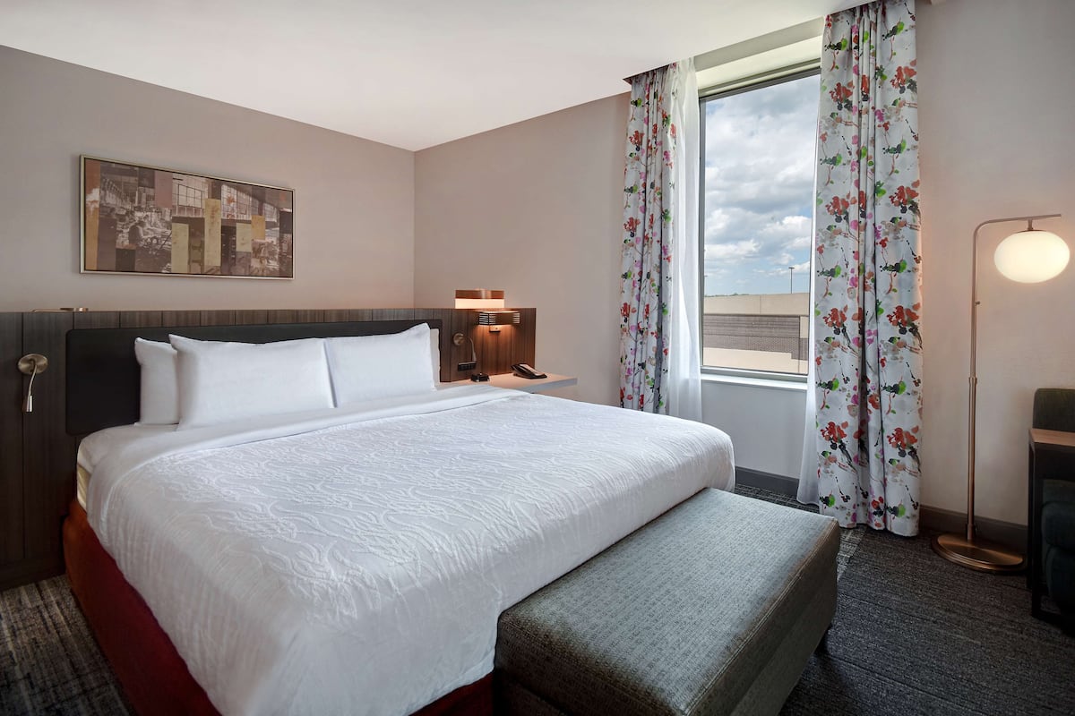 A hotel room with a large bed, a bedside table with a lamp, a window with floral curtains, and a view of the sky.