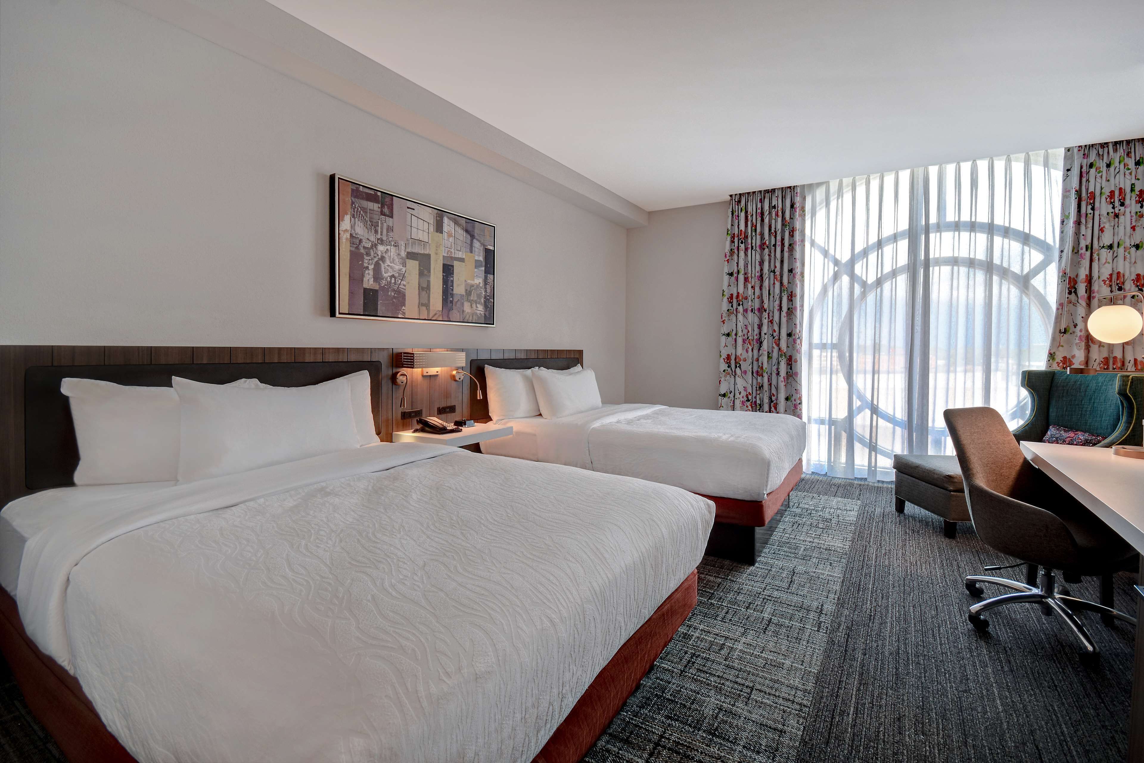 Habitación de hotel con dos camas, un escritorio y un ventanal grande con cortinas.