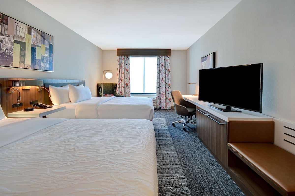 A hotel room with two beds, a desk, a television, and a large window with curtains.
