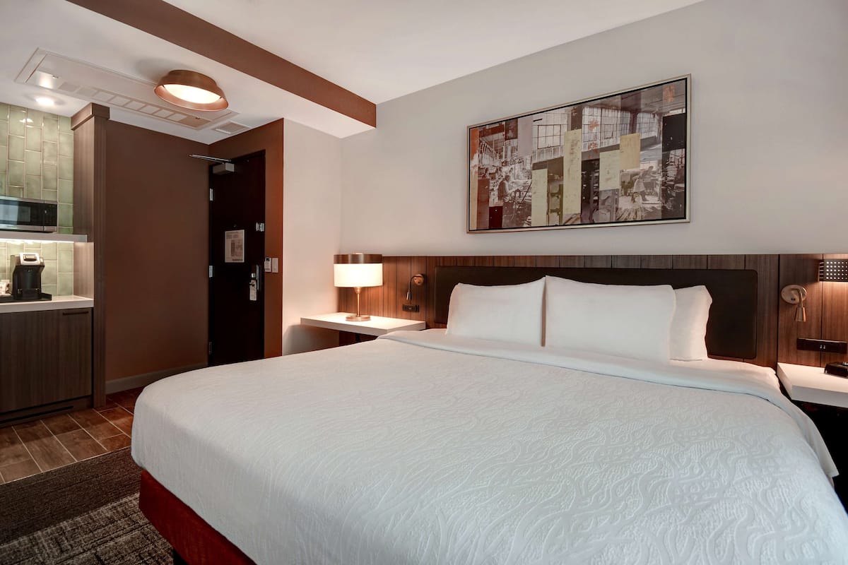 A hotel room with a large bed, a desk with a lamp, a wall-mounted artwork, and a kitchenette area.