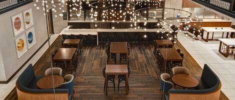 A modern restaurant with multiple seating areas, including round tables and sofas, and a central bar area.