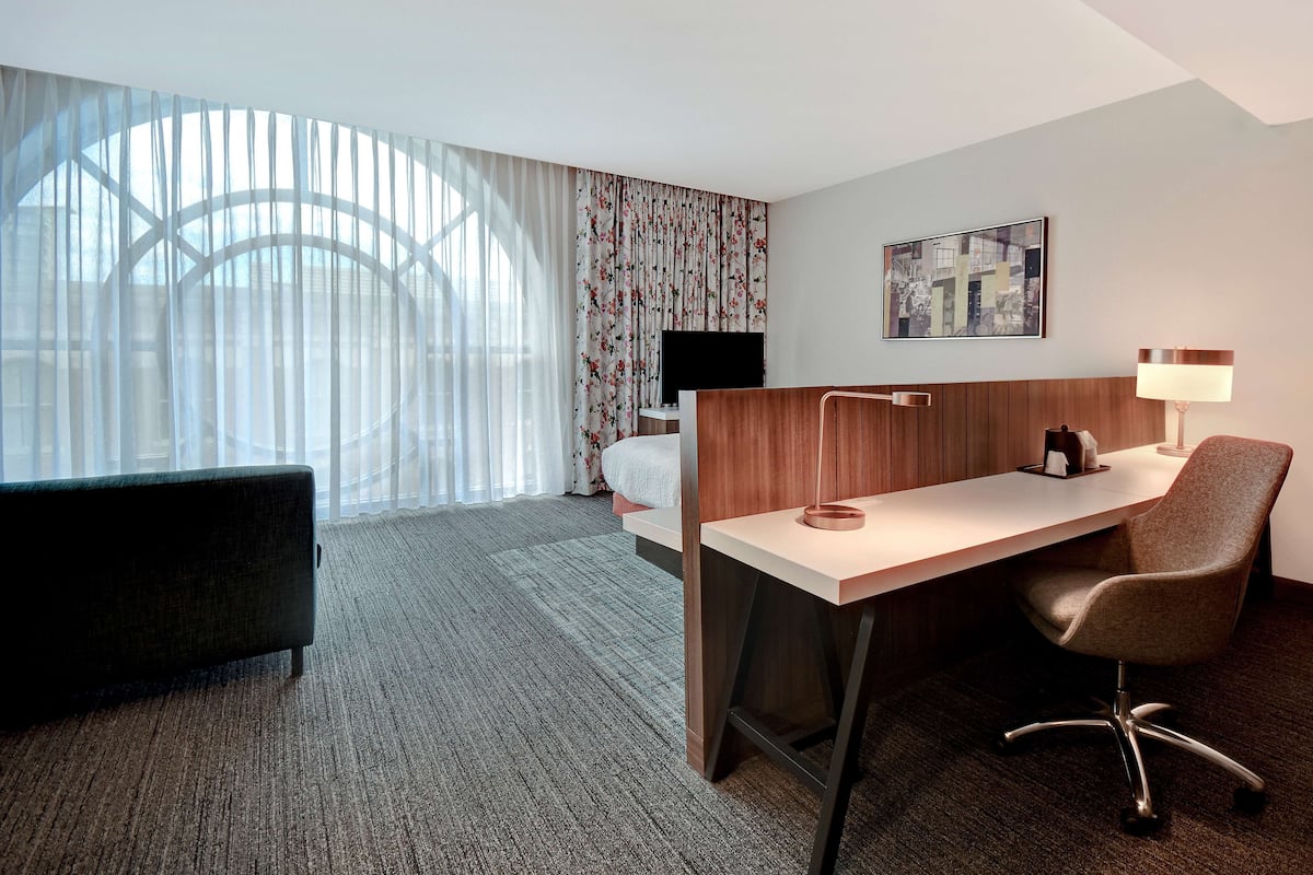 A hotel room with a bed, a desk, a chair, a TV, and a window with sheer curtains.