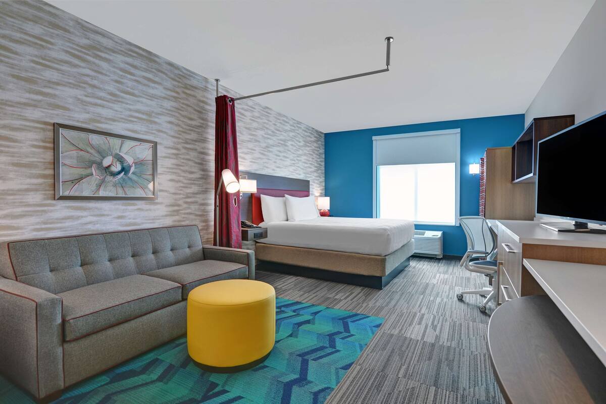 A modern hotel room with a bed, sofa, desk, and a large window.