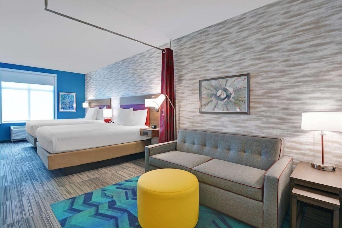 A hotel room with a bed, a sofa, a yellow ottoman, a nightstand, a lamp, and a painting on the wall.