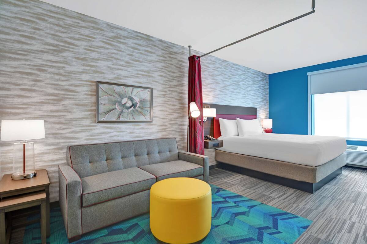 A hotel room with a sofa, a bed, a yellow ottoman, a lamp, a painting, and a window with curtains.