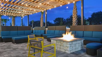 An outdoor seating area with blue sofas and yellow chairs, a stone fire pit, and a pergola with string lights.