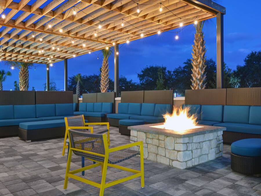 An outdoor seating area with blue sofas and yellow chairs, a stone fire pit, and a pergola with string lights.