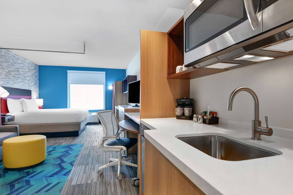 A modern hotel room with a bed, desk, chair, and kitchenette.