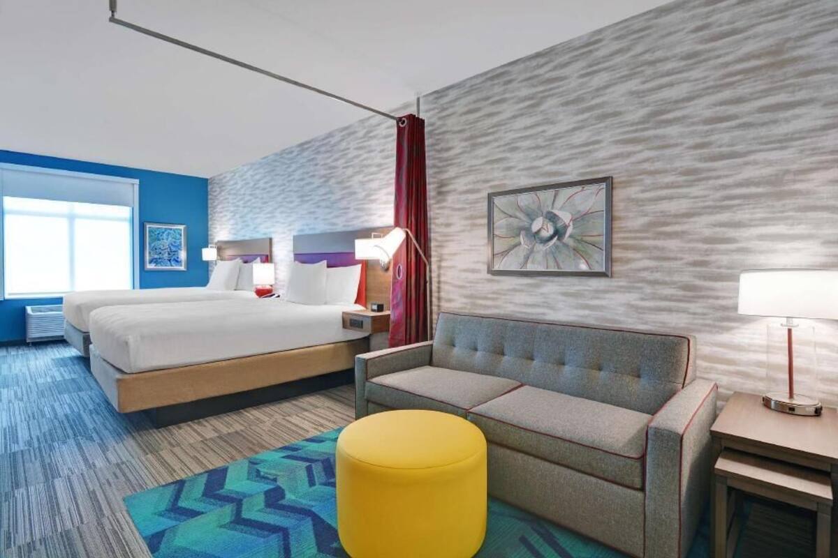 A hotel room with a bed, a sofa, a yellow ottoman, a nightstand, a lamp, and a wall with a textured pattern and a framed picture.