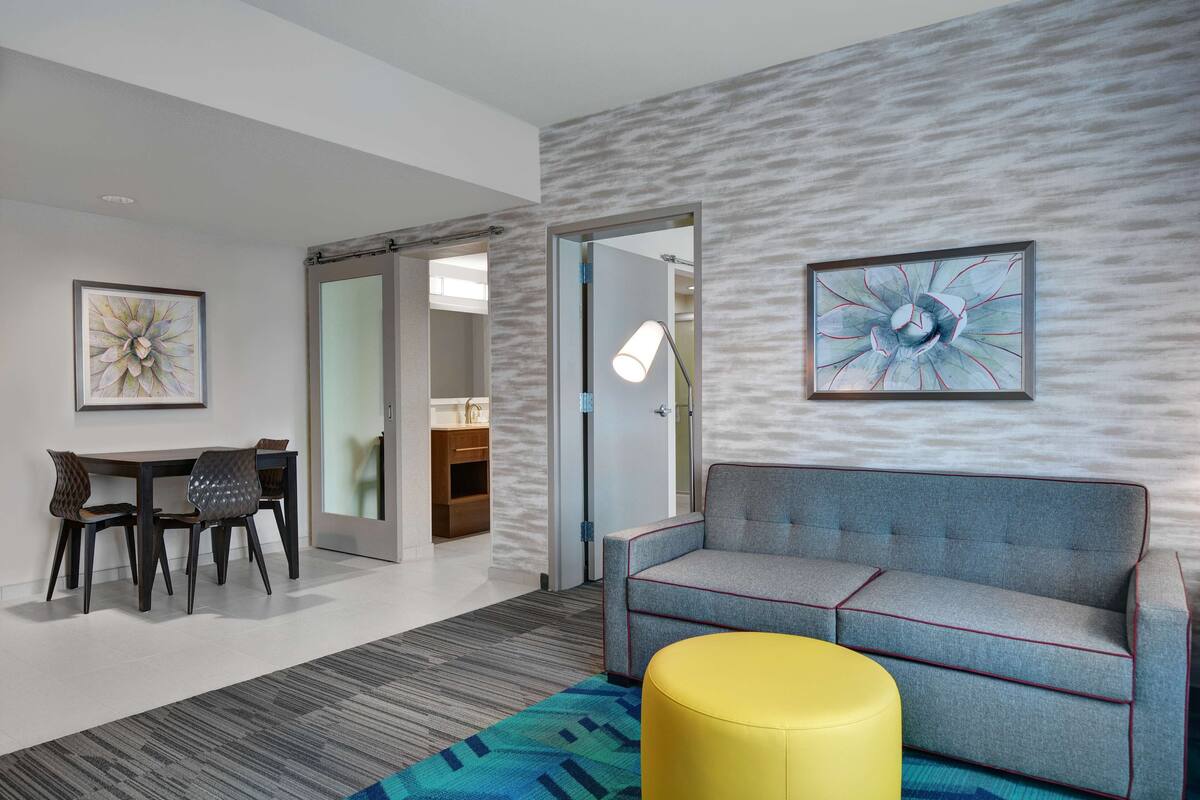 A modern hotel room with a gray sofa, a yellow ottoman, a dining area with a table and chairs, and a large abstract painting on the wall.