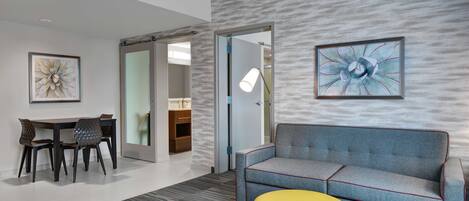 A modern hotel room with a gray sofa, a yellow ottoman, a dining area with a table and chairs, and a large abstract painting on the wall.