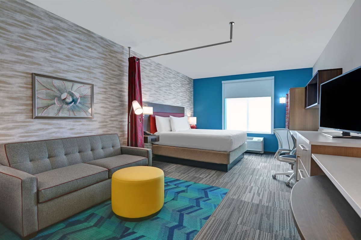 A hotel room with a bed, a sofa, a yellow ottoman, a desk with a TV, and a window with a view.