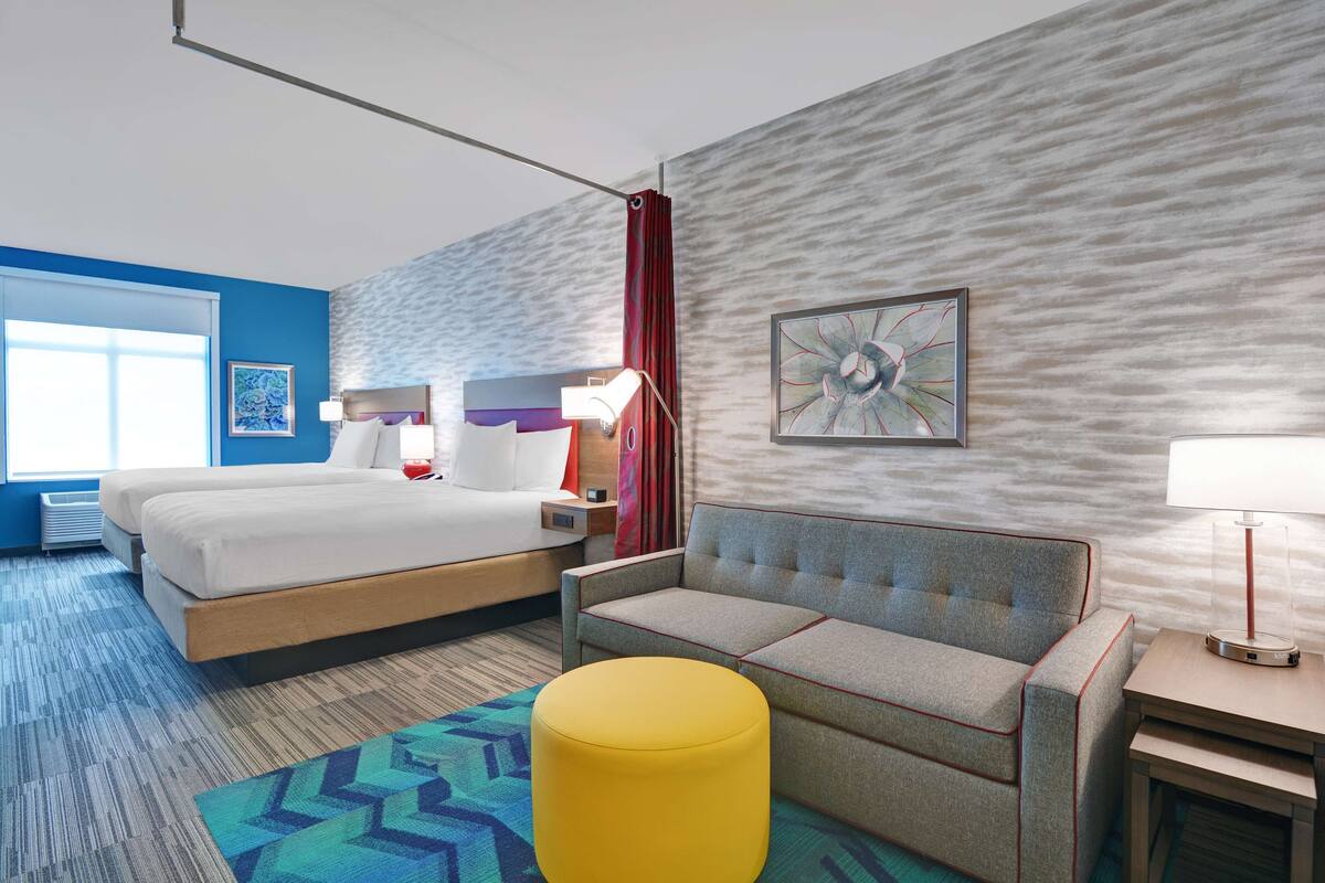 A hotel room with a bed, a sofa, a yellow ottoman, a nightstand, a lamp, and a painting on the wall.