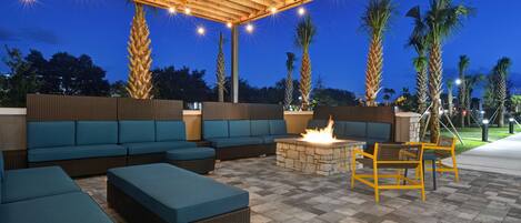 An outdoor patio area with a wooden pergola, blue cushioned seating, a stone fire pit, and palm trees.