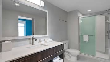 Un baño moderno con un espejo grande, un lavabo blanco, tocador de madera oscura, inodoro blanco y ducha con mampara de vidrio.