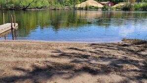 Ein Sandstrand mit einer hölzernen Mole, die in einen ruhigen See führt, umgeben von Grünpflanzen und einigen Gebäuden im Hintergrund.