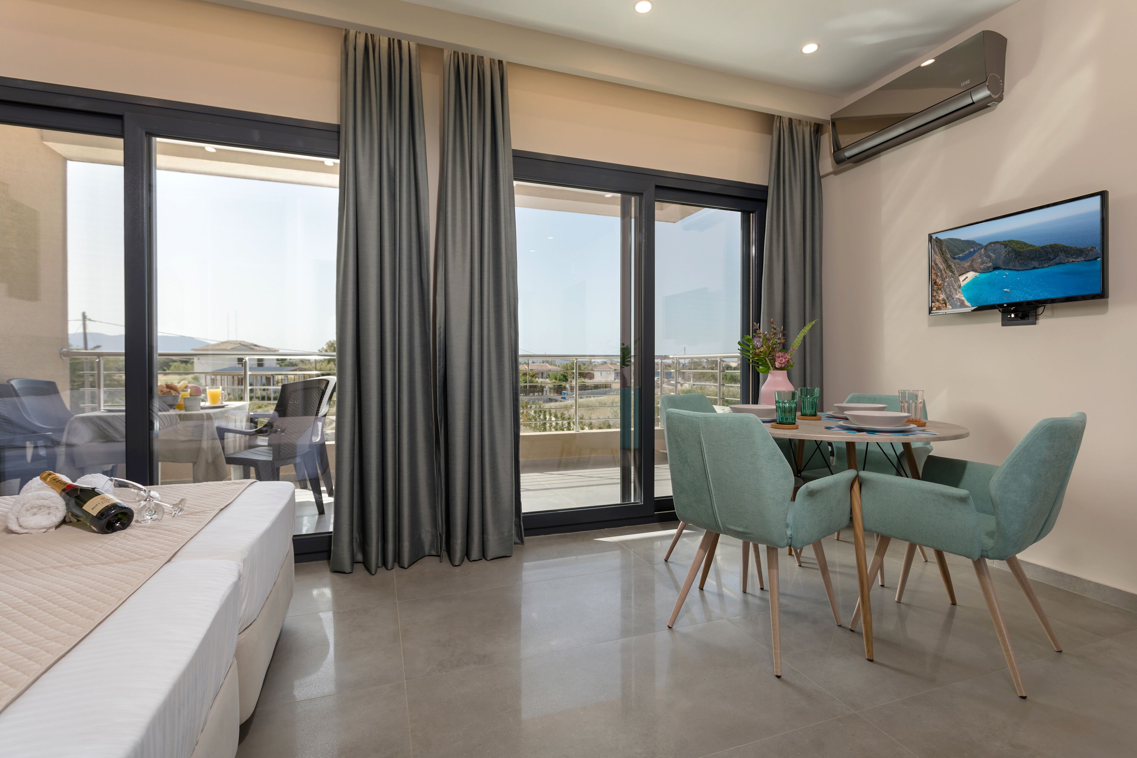 A hotel room with a bed, a balcony with a table and chairs, a TV mounted on the wall, and a large window with curtains.