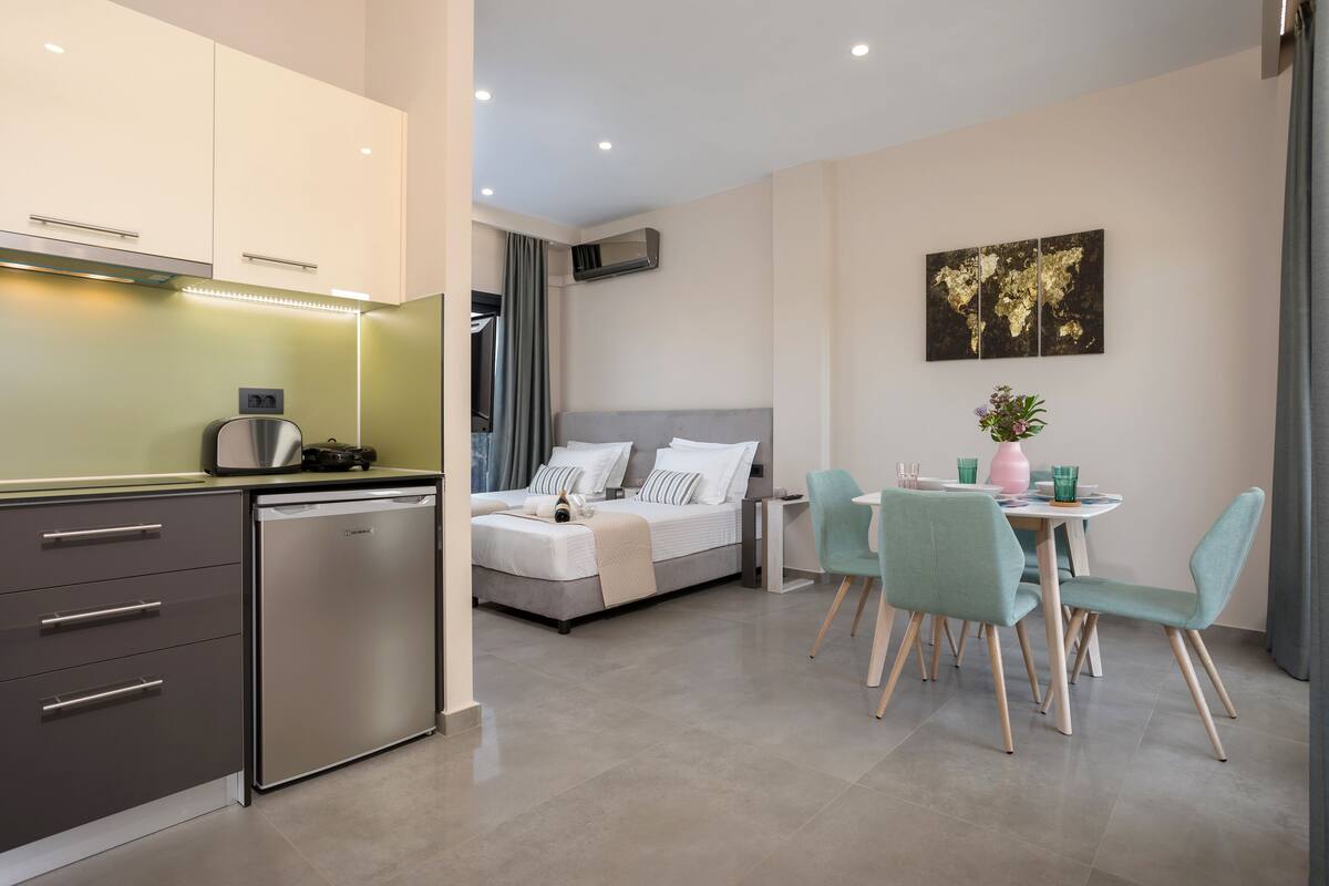 A modern hotel room with a kitchenette, a bed with a grey headboard, a dining area with teal chairs, and a world map artwork on the wall.