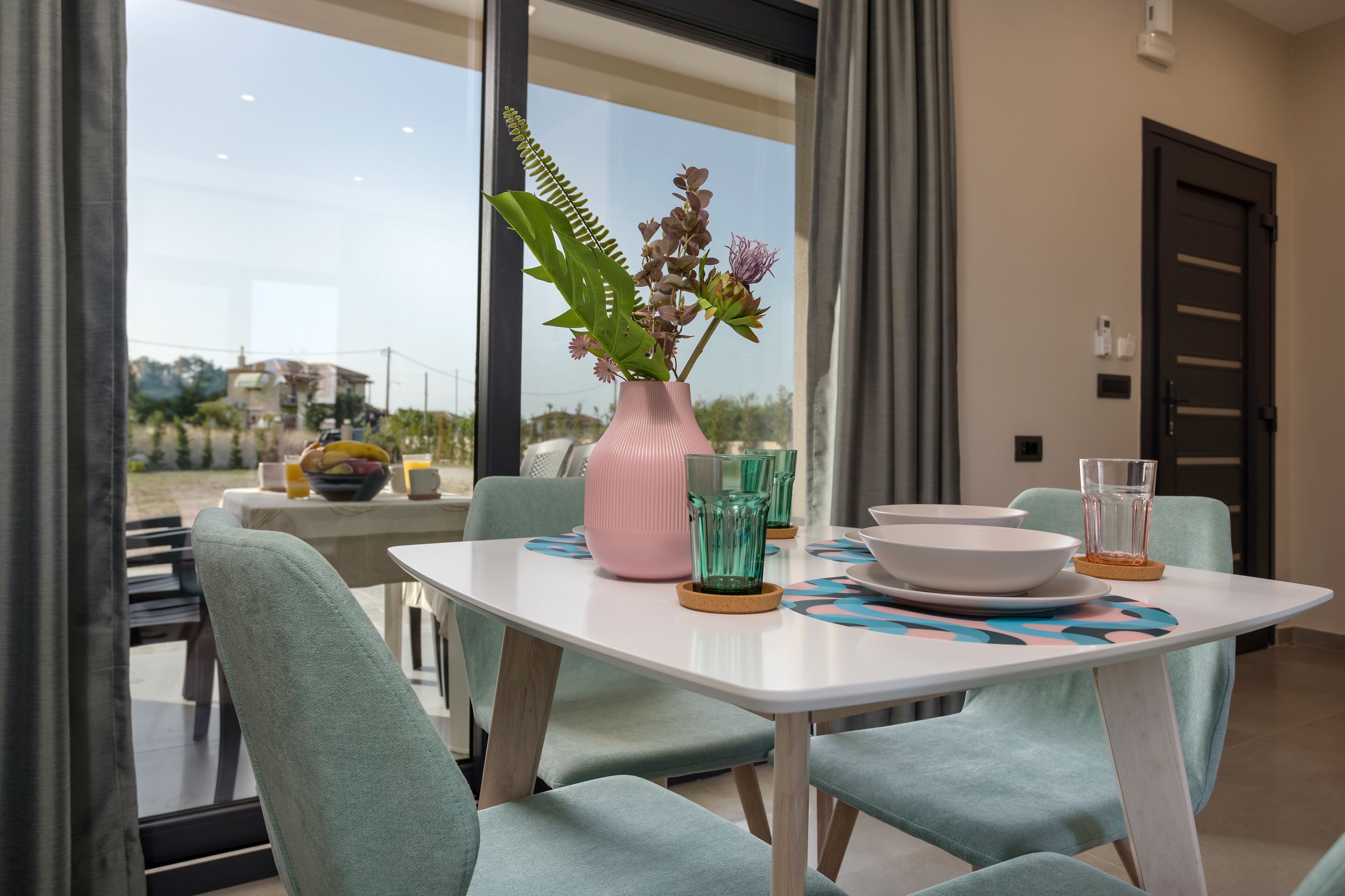 Un coin repas avec une table dressée pour deux personnes, comprenant des chaises turquoise, une table blanche et un vase rose orné de fleurs.