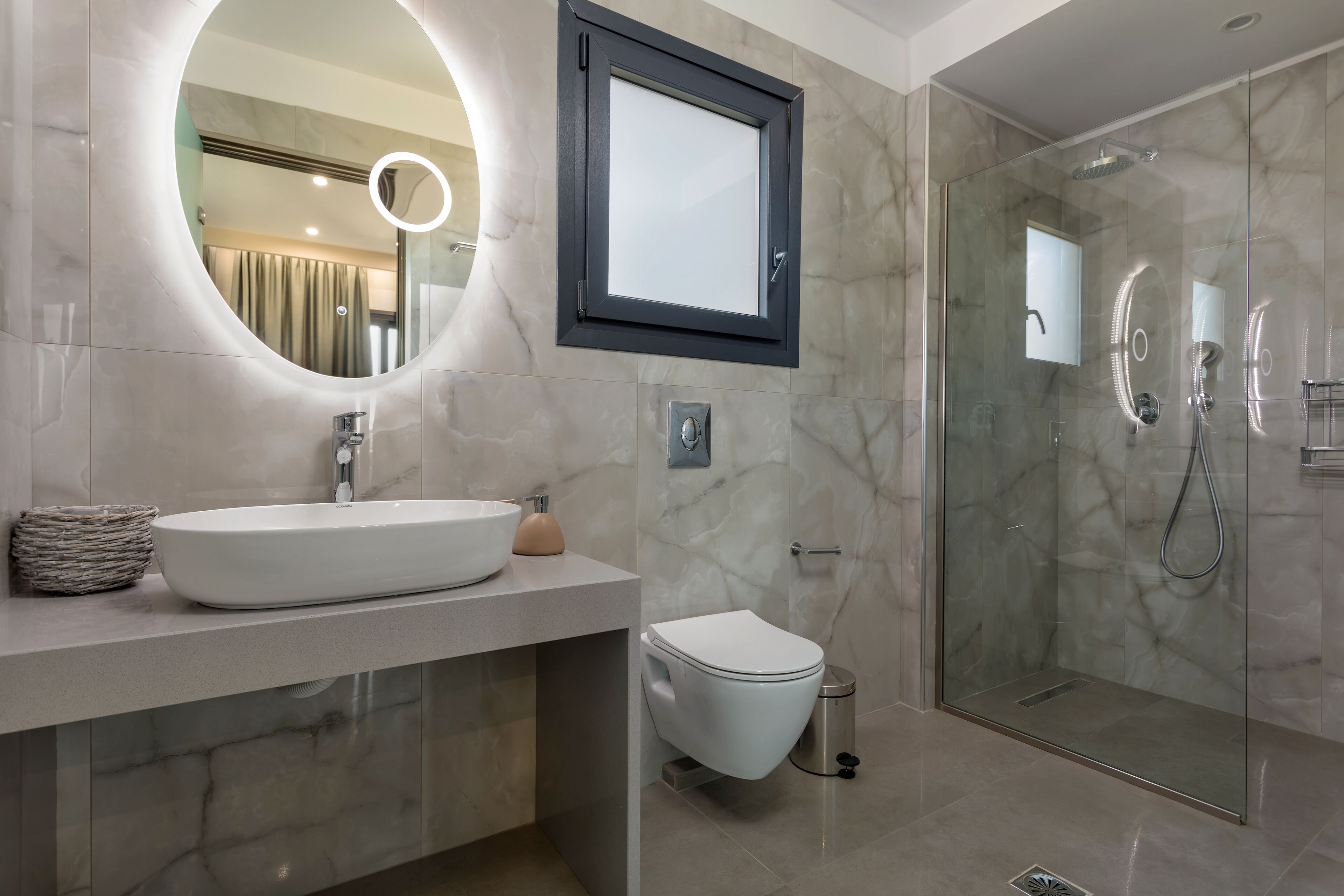 Un baño moderno con un espejo redondo, un lavabo blanco, un inodoro y una zona de ducha con mampara de cristal.