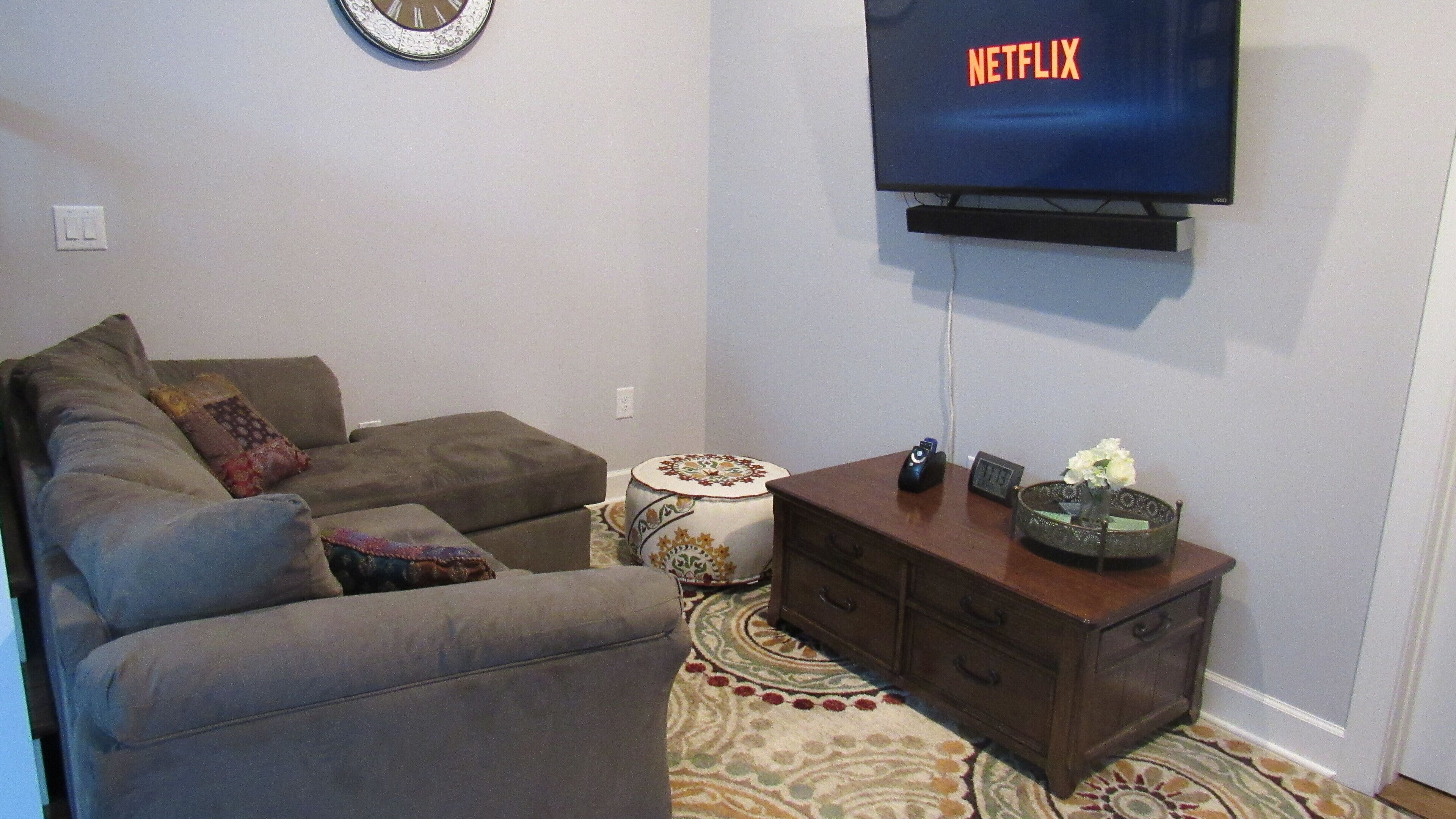 A living room with a sofa, a small round table, a clock, a TV displaying 'NETFLIX', and a decorative vase.
