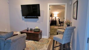 Un salón con un televisor de pantalla plana montado en la pared, una mesa de centro con un reloj y un jarrón de flores, dos sillones y una alfombra estampada.