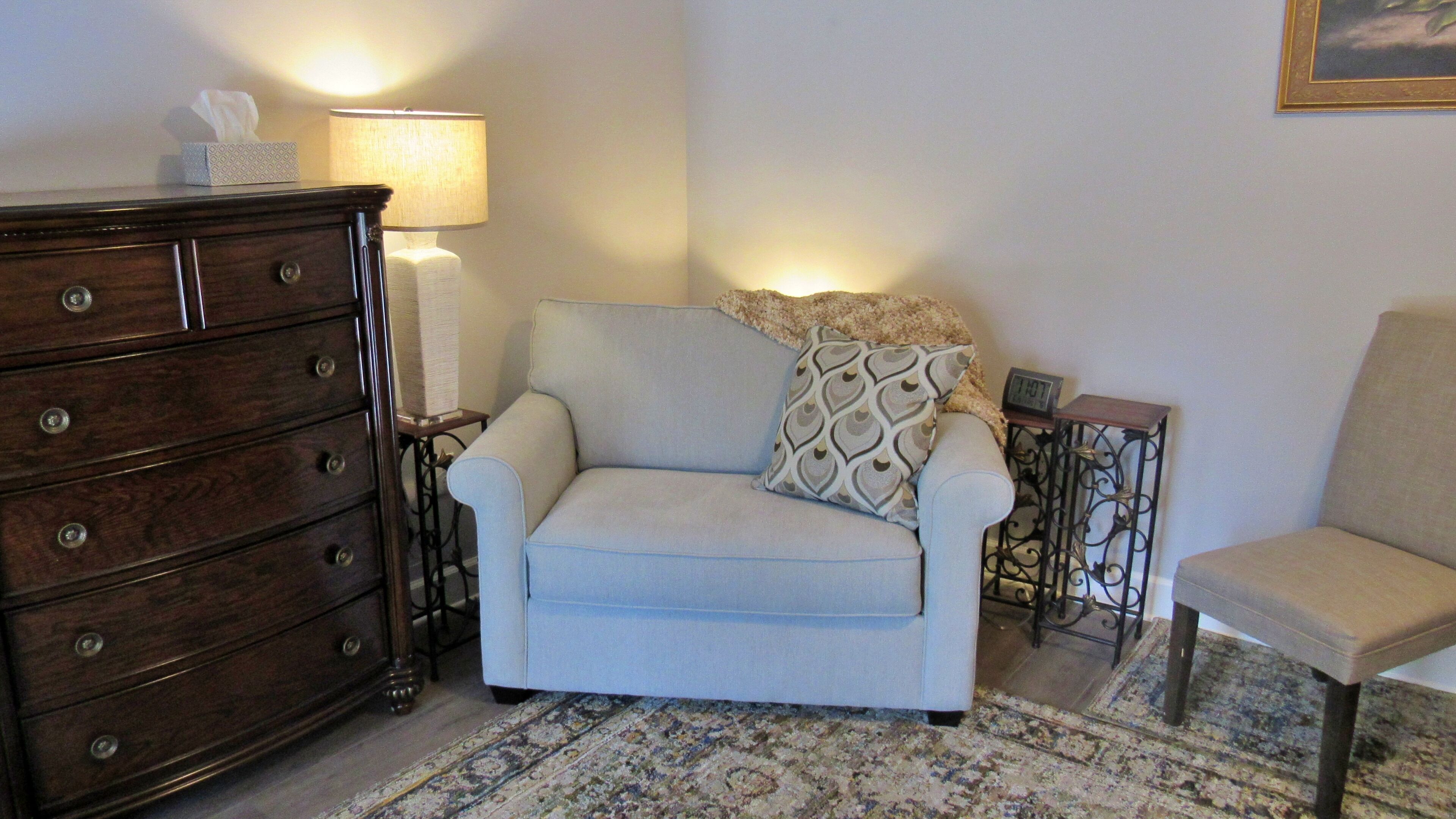A living room with a sofa, a patterned rug, a wooden dresser, a lamp, and a clock on a side table.