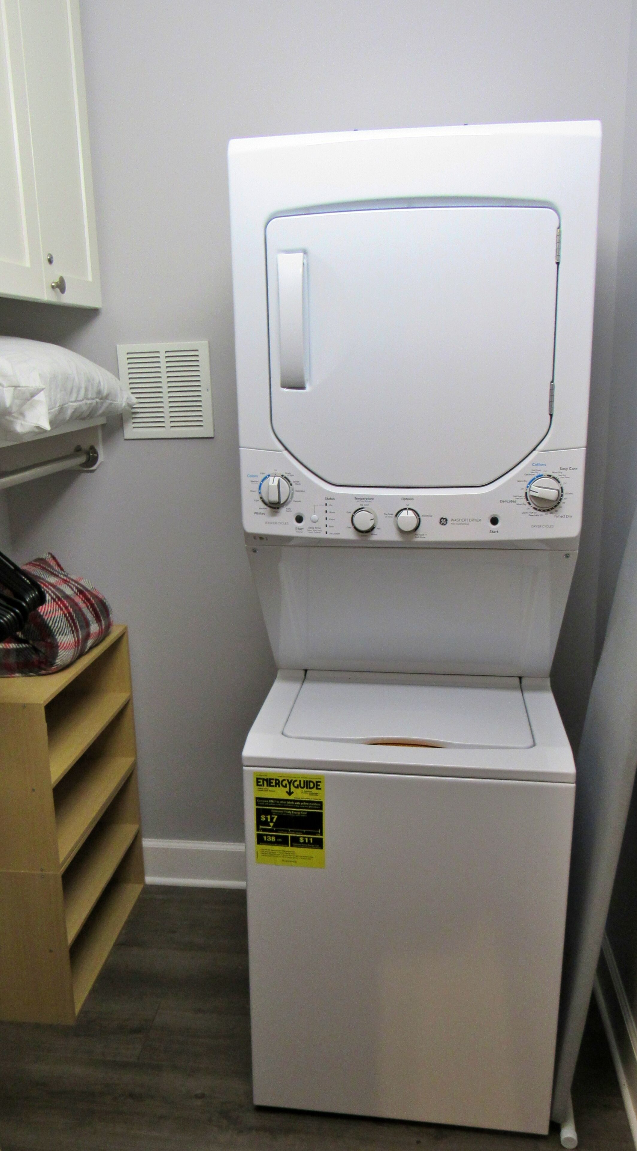 Un appareil combiné lave-linge et sèche-linge à chargement frontal, de couleur blanche, doté d’un panneau de commande, d’une bouche d’aération et d’une étagère en bois sur laquelle est posé un article à motifs écossais.