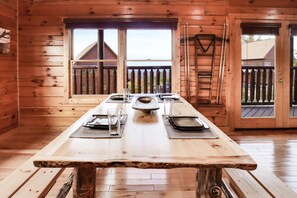 A wooden cabin with a dining table set for four, large windows, and a wooden deck visible through the door.