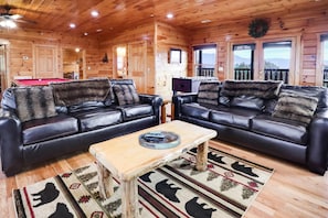 A wooden cabin interior with two leather sofas, a wooden coffee table, and a pool table.