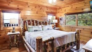 Un dormitorio de cabaña de madera con una cama, dos mesitas de noche y una ventana con vista a árboles.