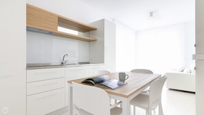 Une cuisine moderne avec des meubles blancs, un plan de travail en bois et un four encastré. On y trouve un coin repas avec une table et des chaises, et un canapé blanc à l’arrière-plan.