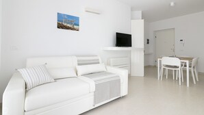 A modern, white-themed living room with a sofa, a flat-screen TV, a dining table with chairs, and a small kitchen area. - Rivazzurra Homes - 37 (Termoli)