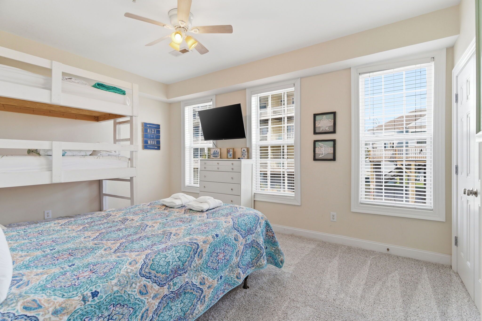 A bedroom with a bunk bed, a flat-screen TV, a bed with a patterned bedspread, a ceiling fan, and a window with blinds.