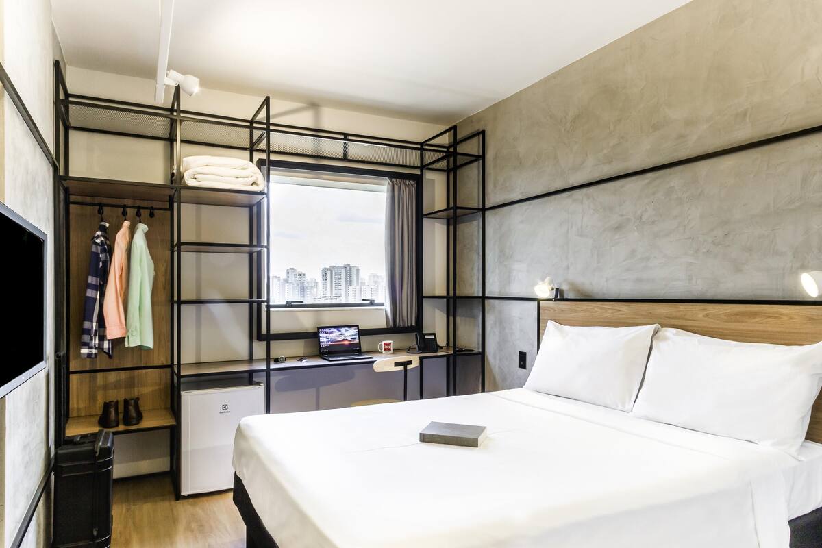 Una habitación de hotel moderna con una cama, un escritorio con una computadora portátil, un armario con ropa colgada y vista a la ciudad por la ventana.