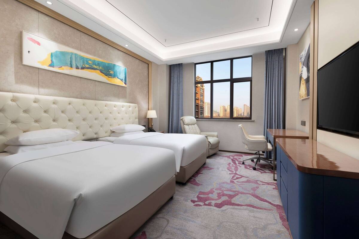 A hotel room with two beds, a desk, a chair, a TV, and a large window with a city view.