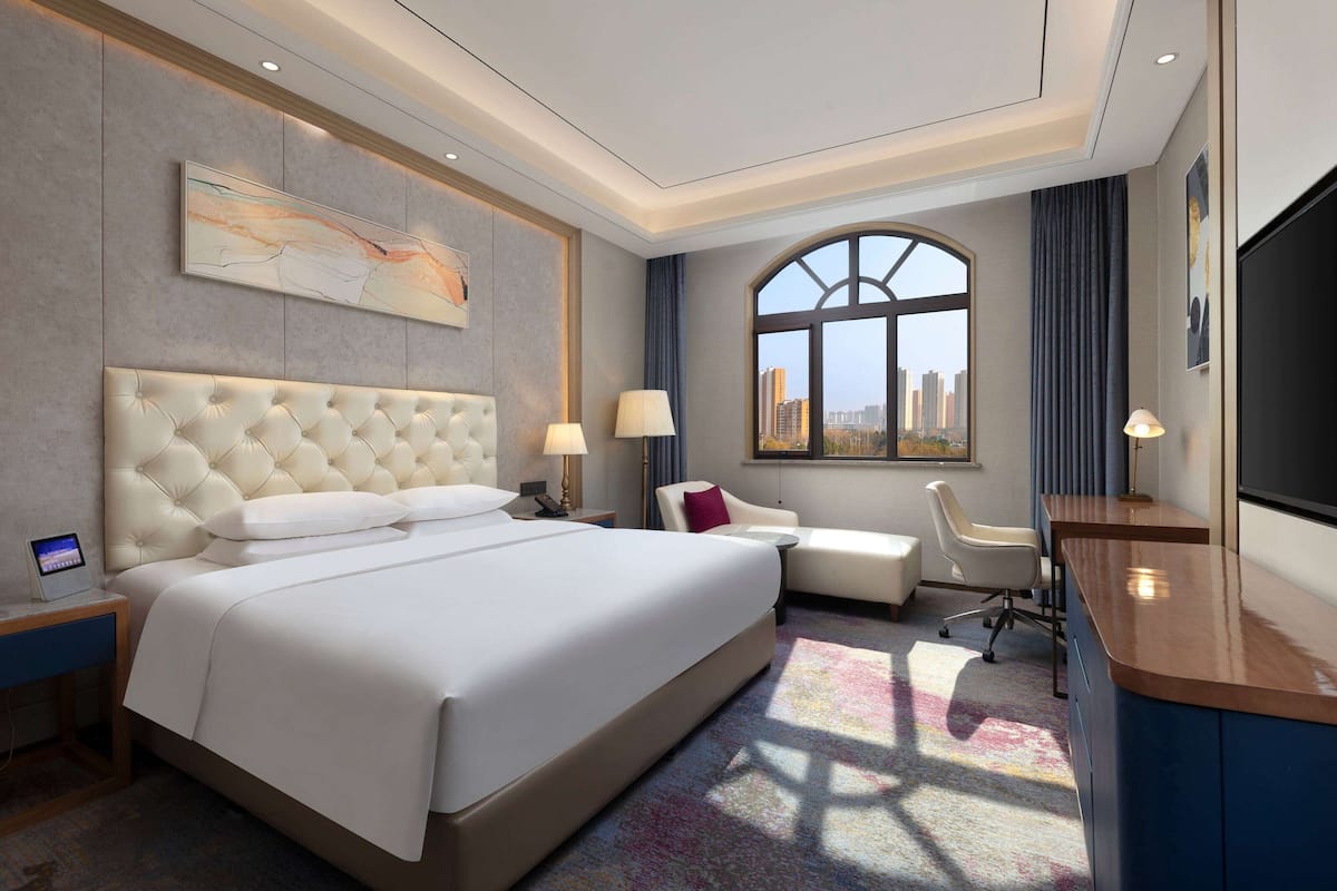 A modern hotel room with a large bed, a desk, a chair, and a view of the cityscape through a large window.