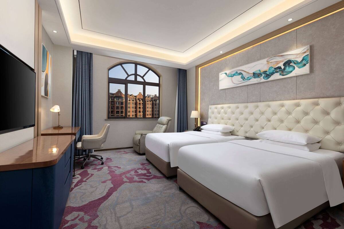 A hotel room with two beds, a desk, a TV, and a large window with a city view.
