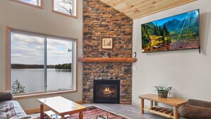 A cozy living room with a stone fireplace, a large window overlooking a lake, a comfortable couch, and a wooden coffee table. - Beautiful Lakeside Home (Woodruff)