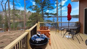 A deck with a table and chairs, a barbecue grill, and a view of a lake surrounded by trees. - Beautiful Lakeside Home (Woodruff)