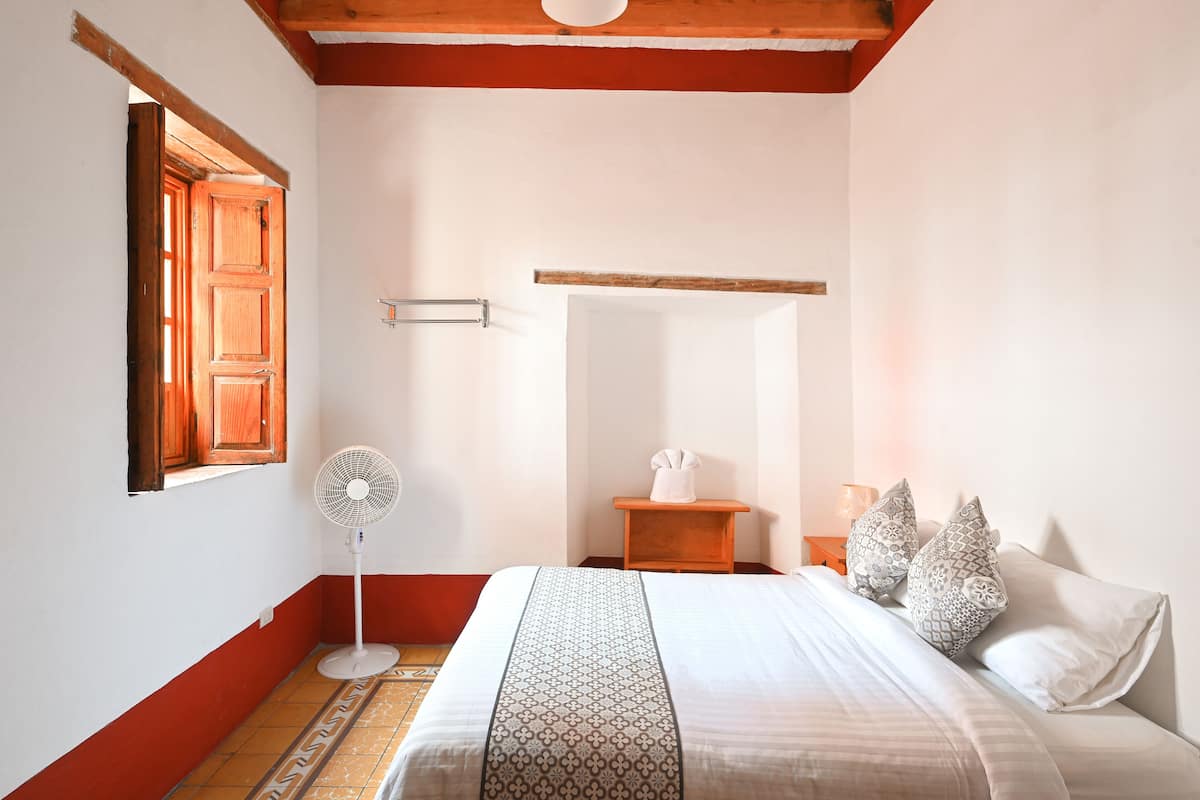 Une chambre à coucher avec un lit, un ventilateur sur pied, une table de chevet avec une lampe et une fenêtre avec des volets en bois.