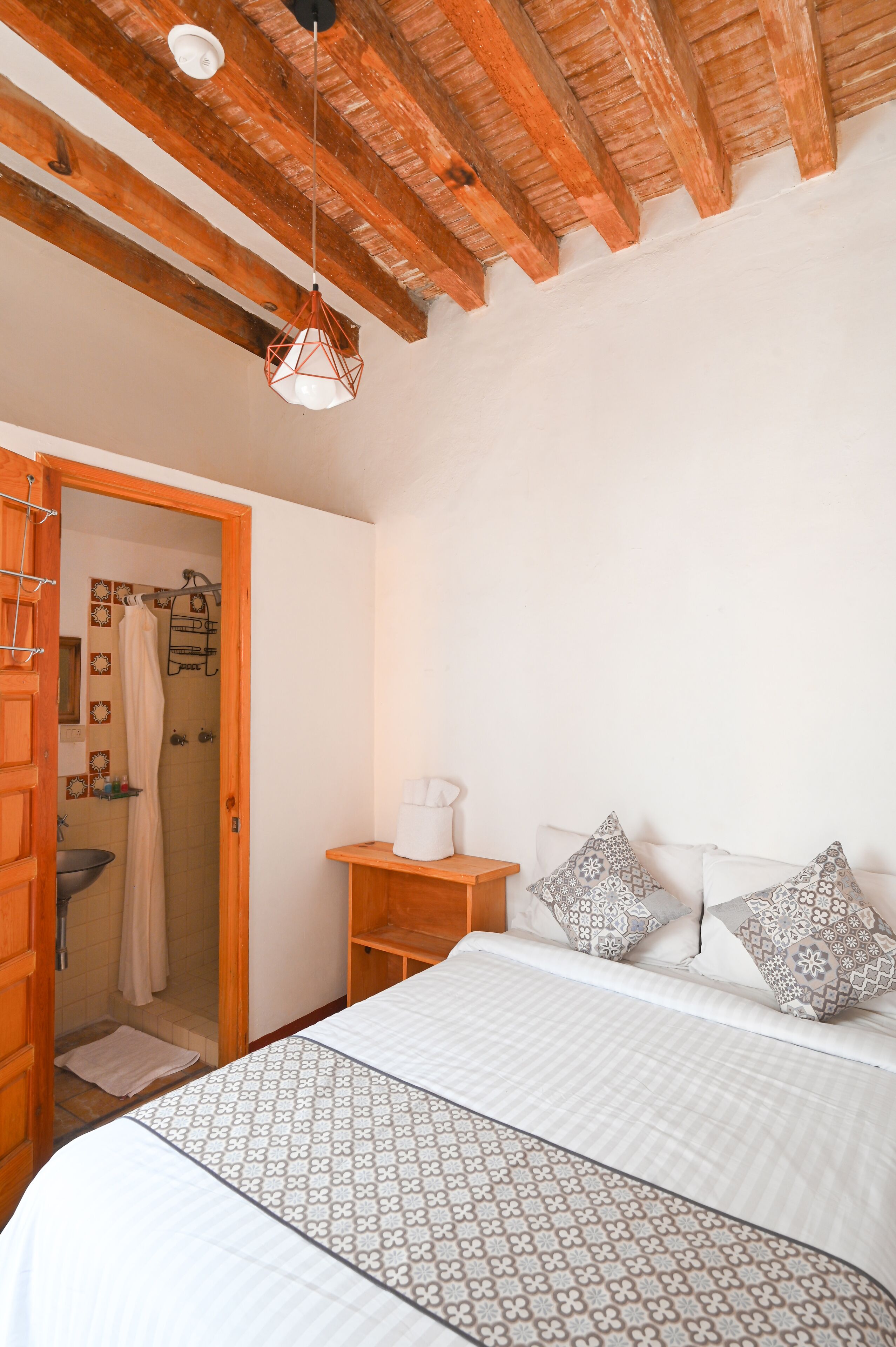 A bedroom with a wooden ceiling, a bed with patterned bedding, a bedside table with a lamp, and a shower area visible through an open door.