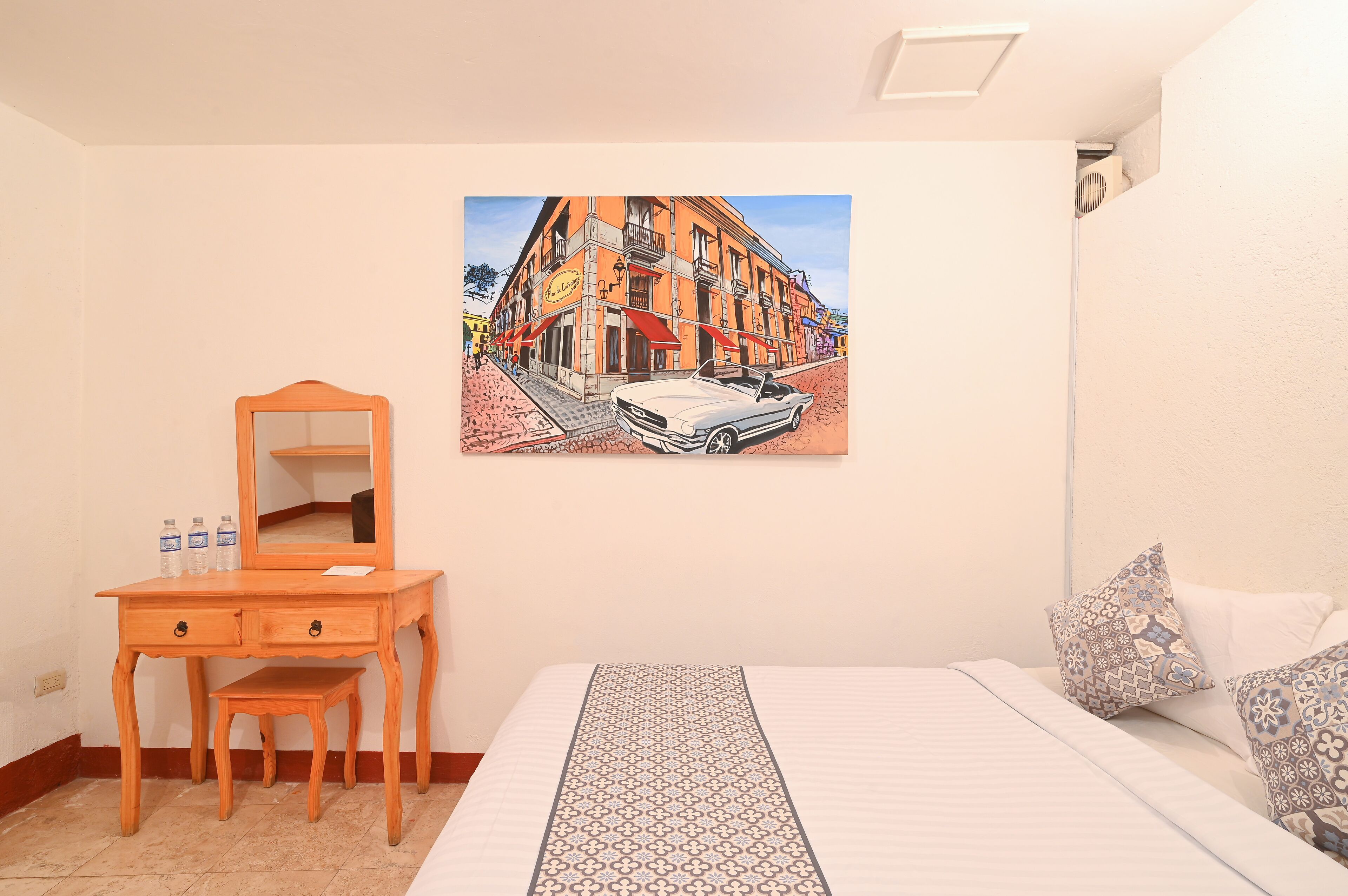 A bedroom with a bed, a wooden dresser, a mirror, and a wall painting of a street scene.