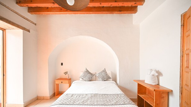 A bedroom with a bed, a nightstand, a wooden bench, and a white wall with an arched doorway. - Hotel Mesón Cuévano (Guanajuato)