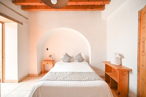 A bedroom with a bed, a nightstand, a wooden bench, and a white wall with an arched doorway. - Hotel Mesón Cuévano (Guanajuato)