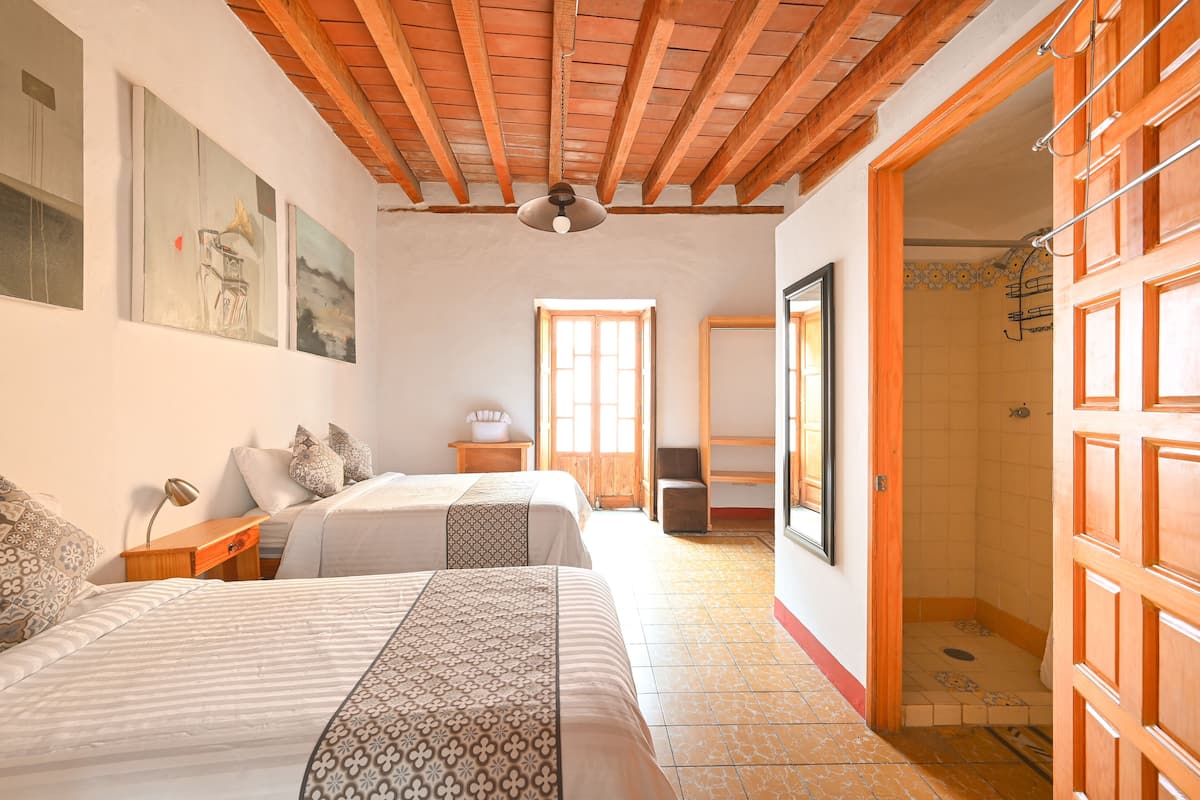 Une chambre avec deux lits, des poutres apparentes en bois au plafond, une fenêtre et une salle de bain avec douche.