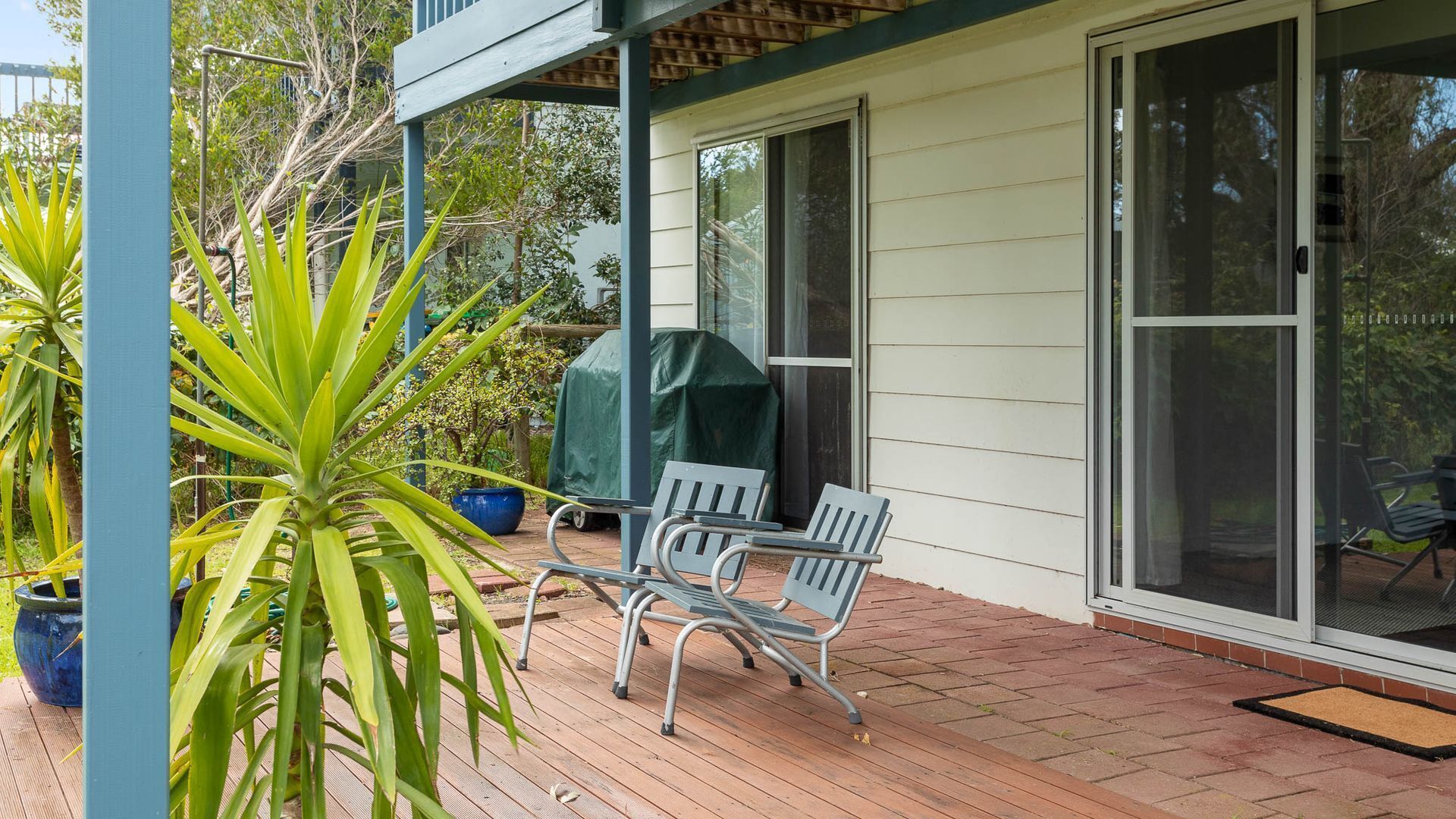 Eine Terrasse mit zwei Metallstühlen, einem Tisch, einem Grill mit Überdachung und einem großen Kübelpflanzen.